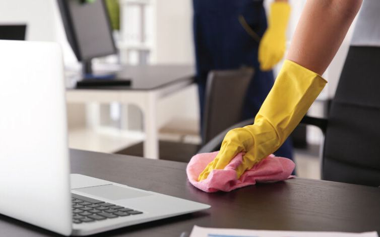 Cleaner in a yellow glove wiping an office desk with a pink microfibre cloth beside a laptop