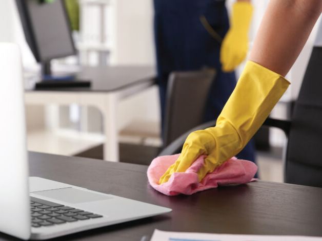 Cleaner in a yellow glove wiping an office desk with a pink microfibre cloth beside a laptop
