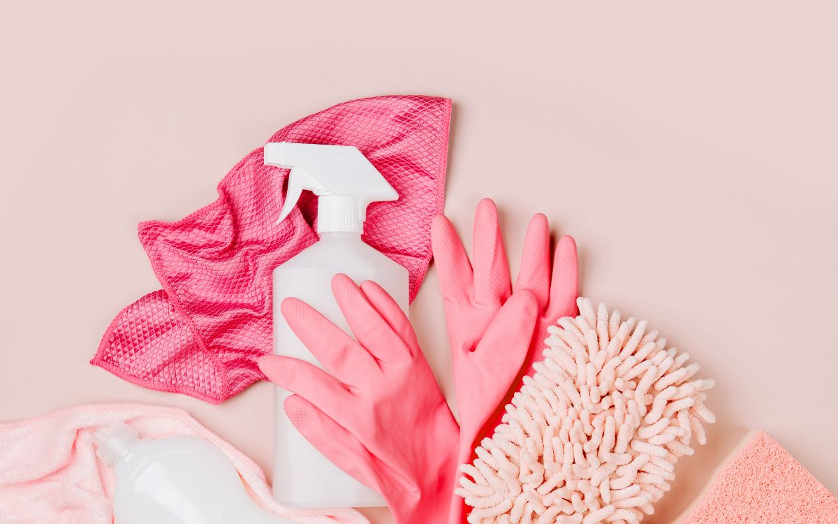 Spray bottle, rubber gloves, sponge and microfibre cloths laid out on a pink background