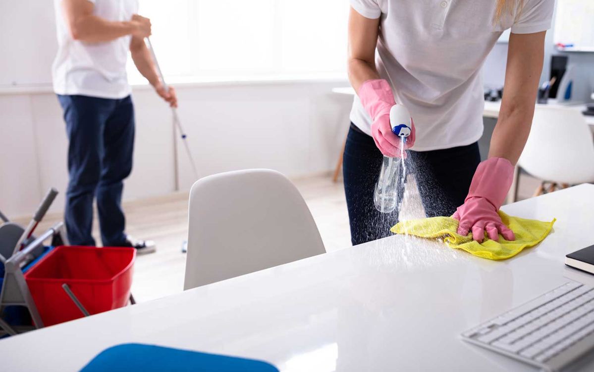 Two cleaners working an office together, one mopping the floor and one spraying and wiping a white table