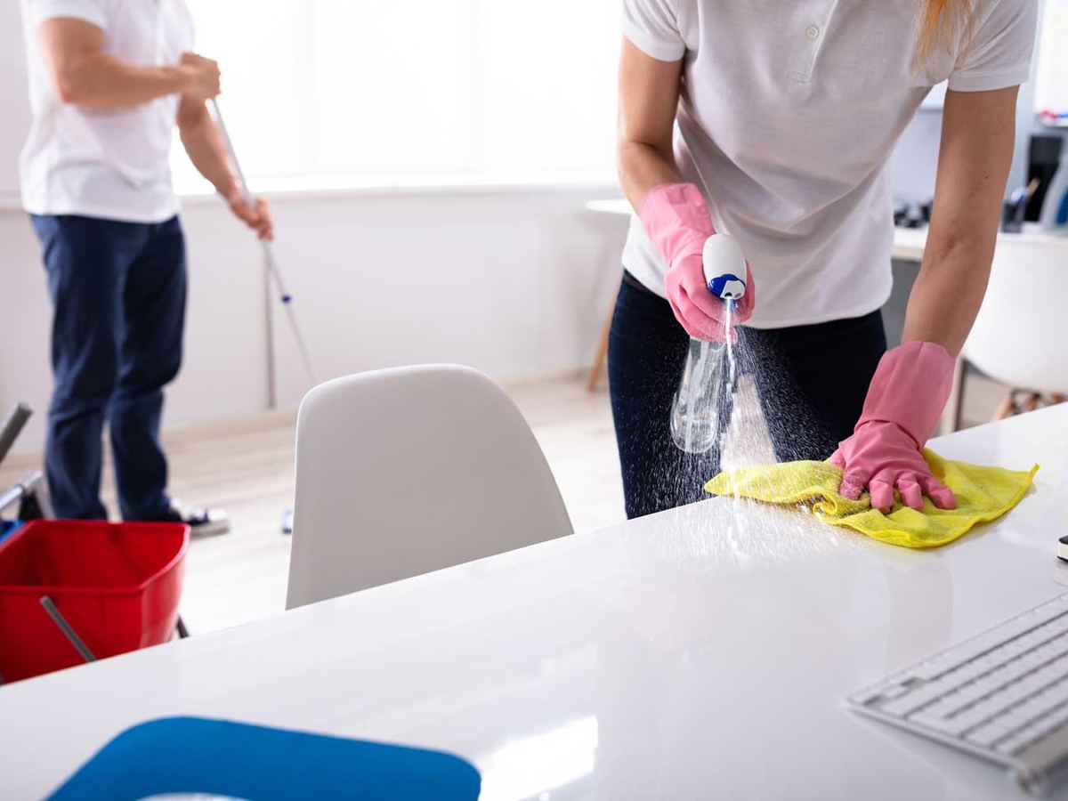 Two cleaners working an office together, one mopping the floor and one spraying and wiping a white table