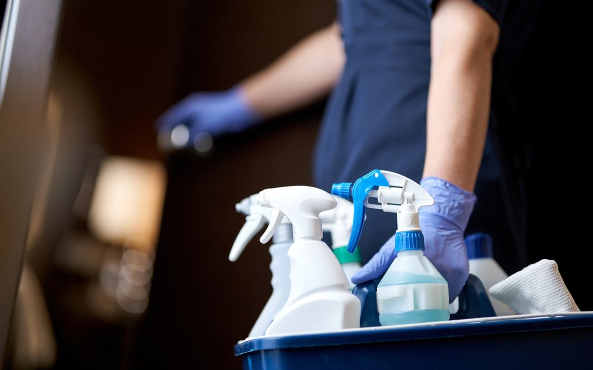 Cleaner in a navy uniform carrying a tray of spray bottles along a corridor