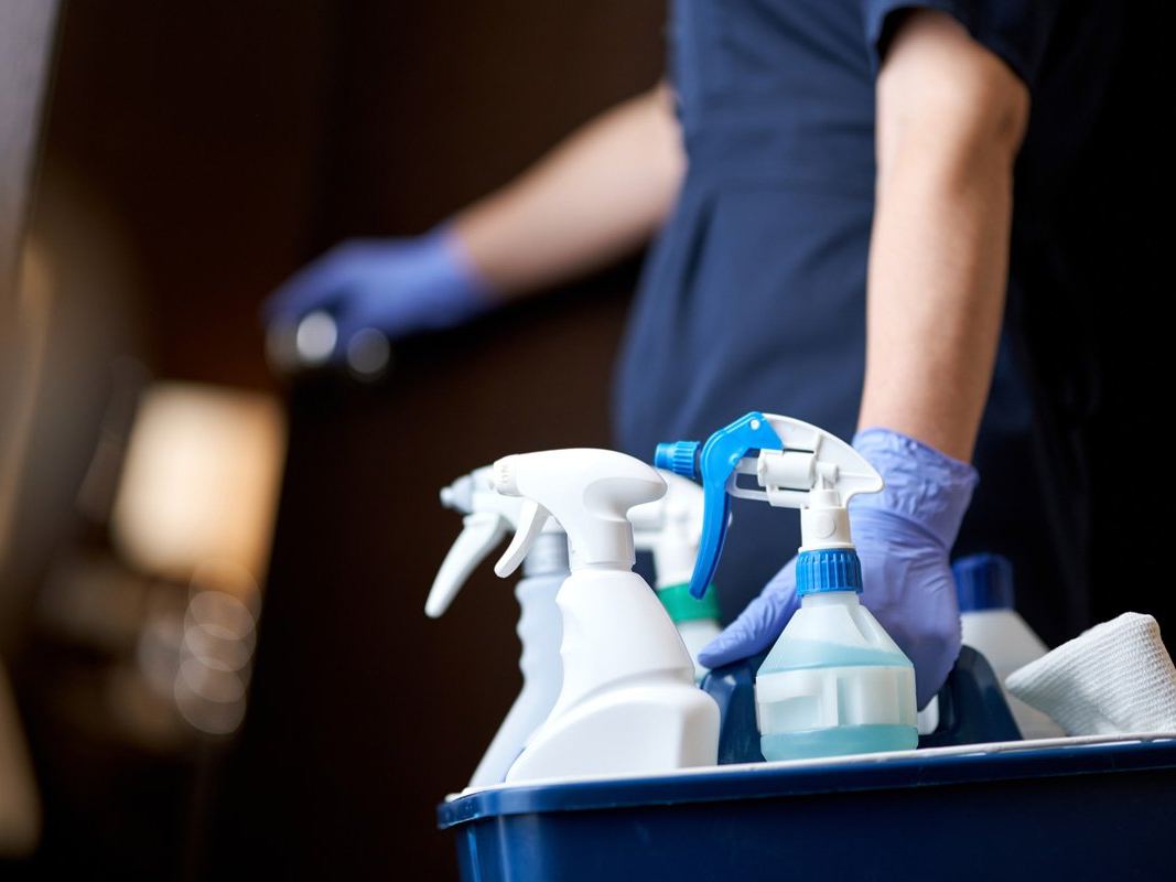 Cleaner in a navy uniform carrying a tray of spray bottles along a corridor